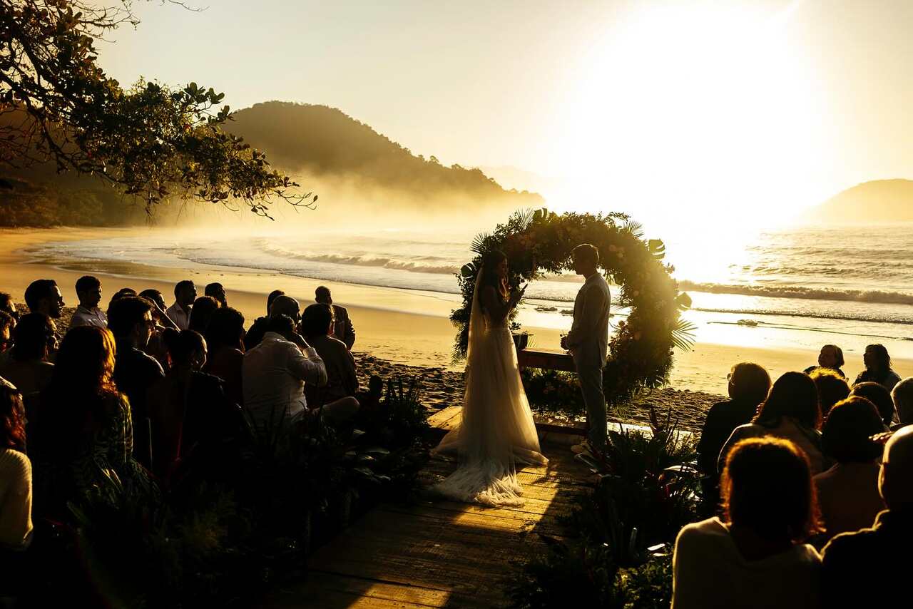 Casamento ao ar livre na praia, com o casal trocando votos sob um arco floral e os convidados assistindo, enquanto o sol nasce ao fundo.