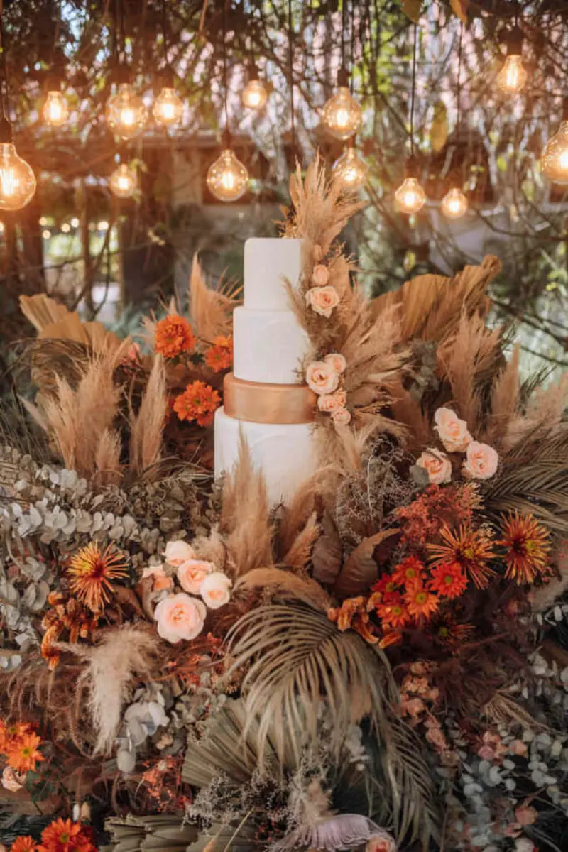 bolo de quatro andares branco minimalista. Em mesa decorada boho chic focada em elementos naturais e tons quente