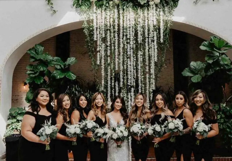 mulheres sorrindo em um evento formal, com uma noiva ao centro usando um vestido branco e as damas de honra ao redor vestindo preto e segurando buquês de flores brancas