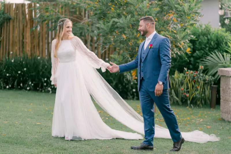 casal caminhando juntos em um cenário de casamento ao ar livre, com a noiva vestindo um vestido de noiva longo com mangas compridas e uma longa cauda, e o noivo usando um terno azul.