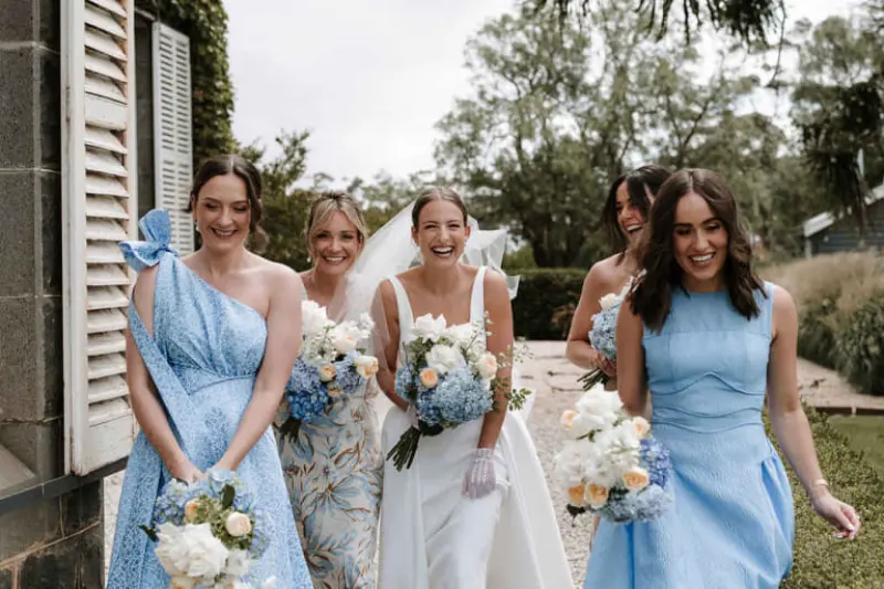 Esta imagem mostra madrinhas de casamento com vestidos azuis e brancos, com destaque para a noiva no centro segurando um buqu&ecirc; branco e azul
