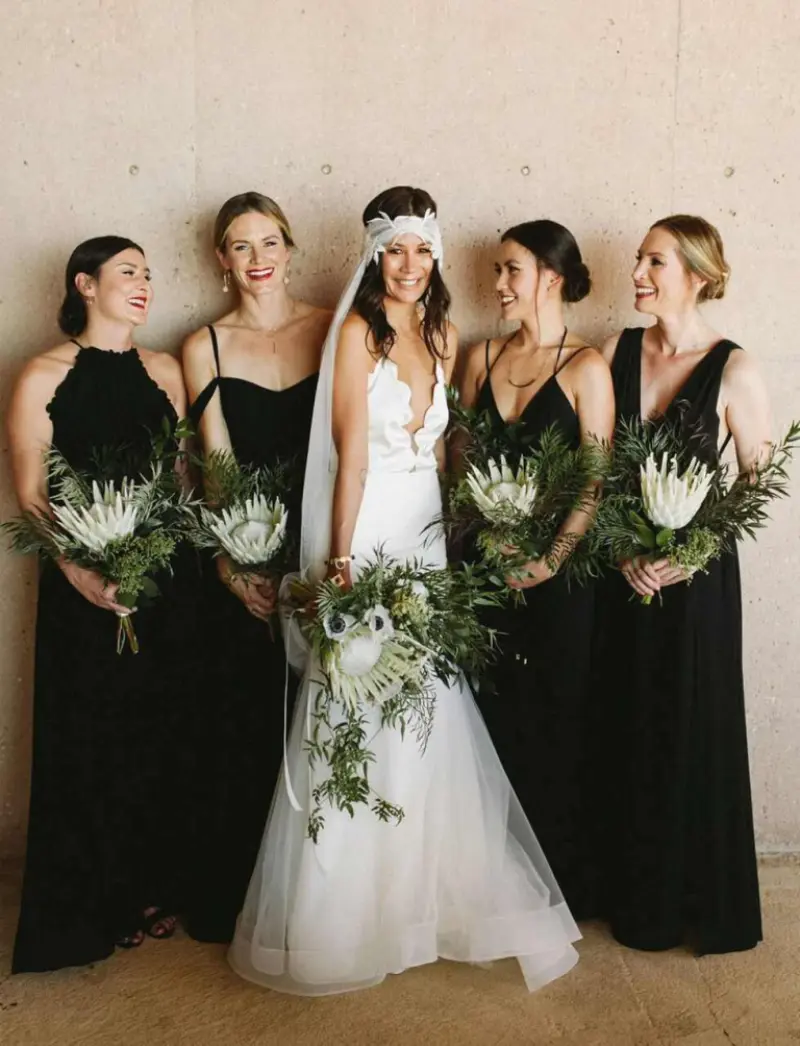 noiva em um vestido branco de um ombro só ao lado de duas madrinhas vestindo vestidos pretos de um ombro só, todos segurando buquês com flores vermelhas e cor-de-rosa