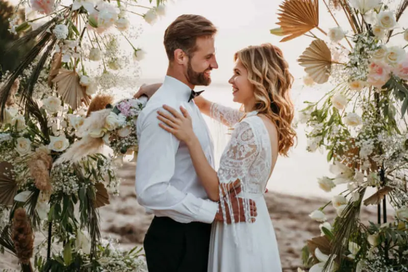 casal em trajes de casamento, posando em frente a um arco floral em um cen&aacute;rio de praia