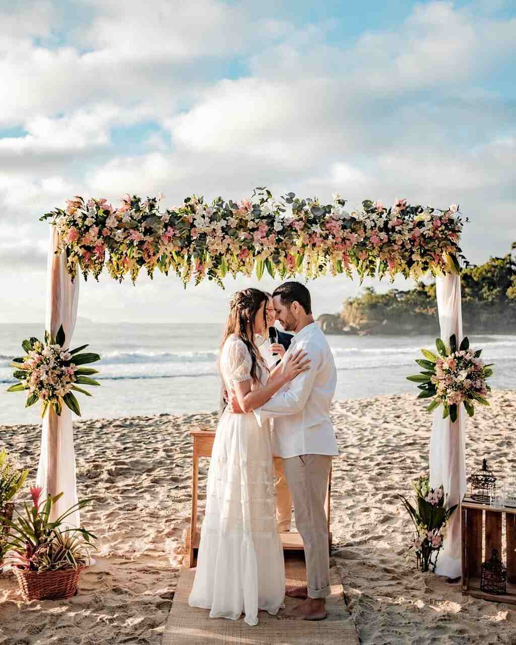 Casal trocando votos durante a cerimônia de casamento na praia, sob um arco floral, com o mar ao fundo e o céu claro.