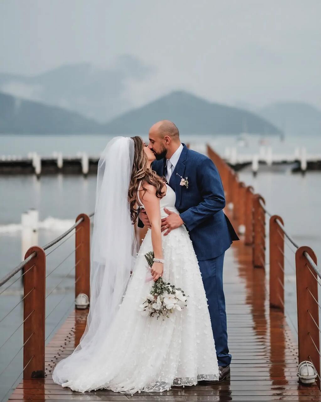 Casal se beijando no meio de um pier, com a noiva usando um vestido de noiva branco e o noivo em um terno azul, com montanhas e o mar ao fundo.