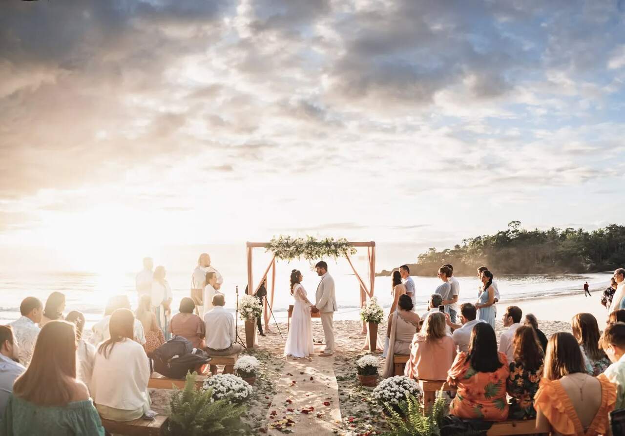 Casamento ao ar livre na praia durante o pôr do sol, com o casal trocando votos sob um arco floral e os convidados assistindo sentados em bancos, com a praia ao fundo.