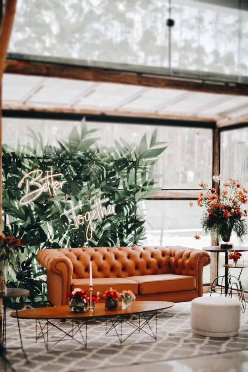 ambiente de decora&ccedil;&atilde;o no estilo Boho Chic, caracterizado pela mistura de texturas naturais, tons terrosos e plantas, como o sof&aacute; de couro estilo Chesterfield e o painel de folhagens.&nbsp;