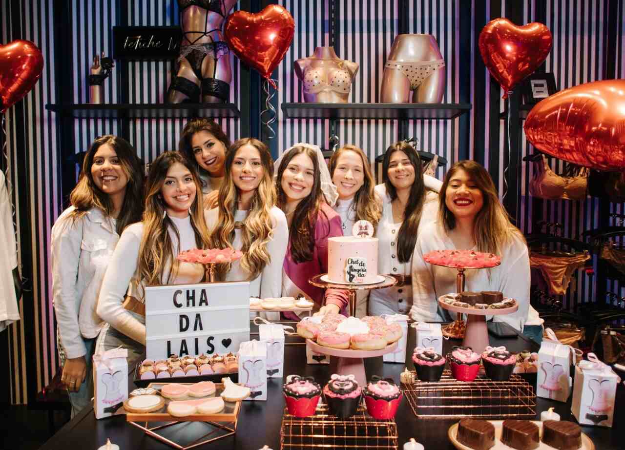 Grupo de mulheres sorrindo durante um "Chá de Lingerie", com decoração temática e doces personalizados como cupcakes e bolos, ao fundo há balões em forma de coração.
