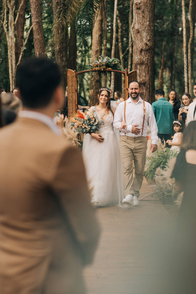 Noiva caminhando com seu pai pelo corredor de casamento ao ar livre, com natureza ao fundo.