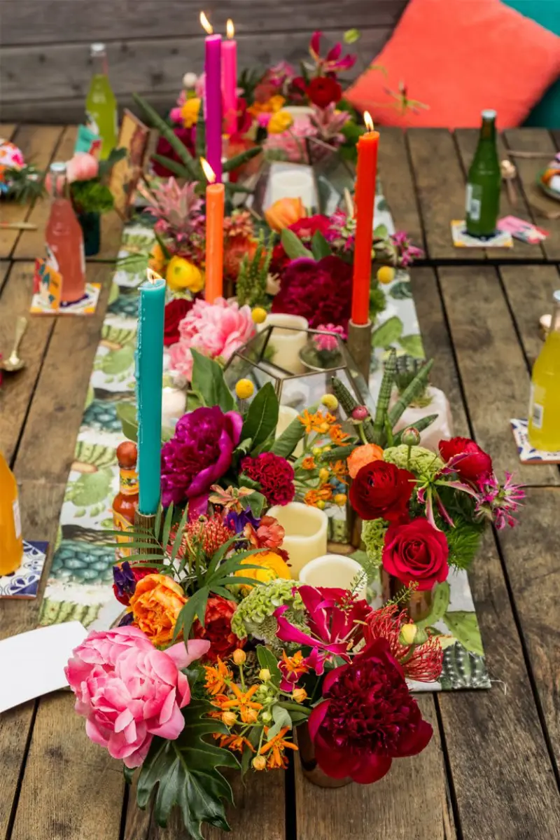 caminho de mesa r&uacute;stica com velas coloridas e arranjo floral vibrante em tons de rosa e laranja.