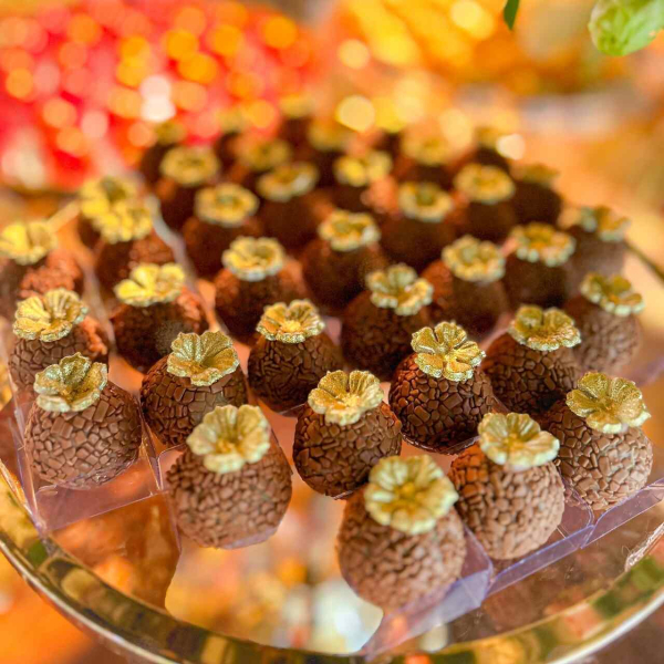 capa post: Brigadeiros de chocolate com cobertura dourada e florzinhas douradas no topo, decorando uma mesa de doces com cores quentes e brilhantes. A imagem tem um fundo difuso