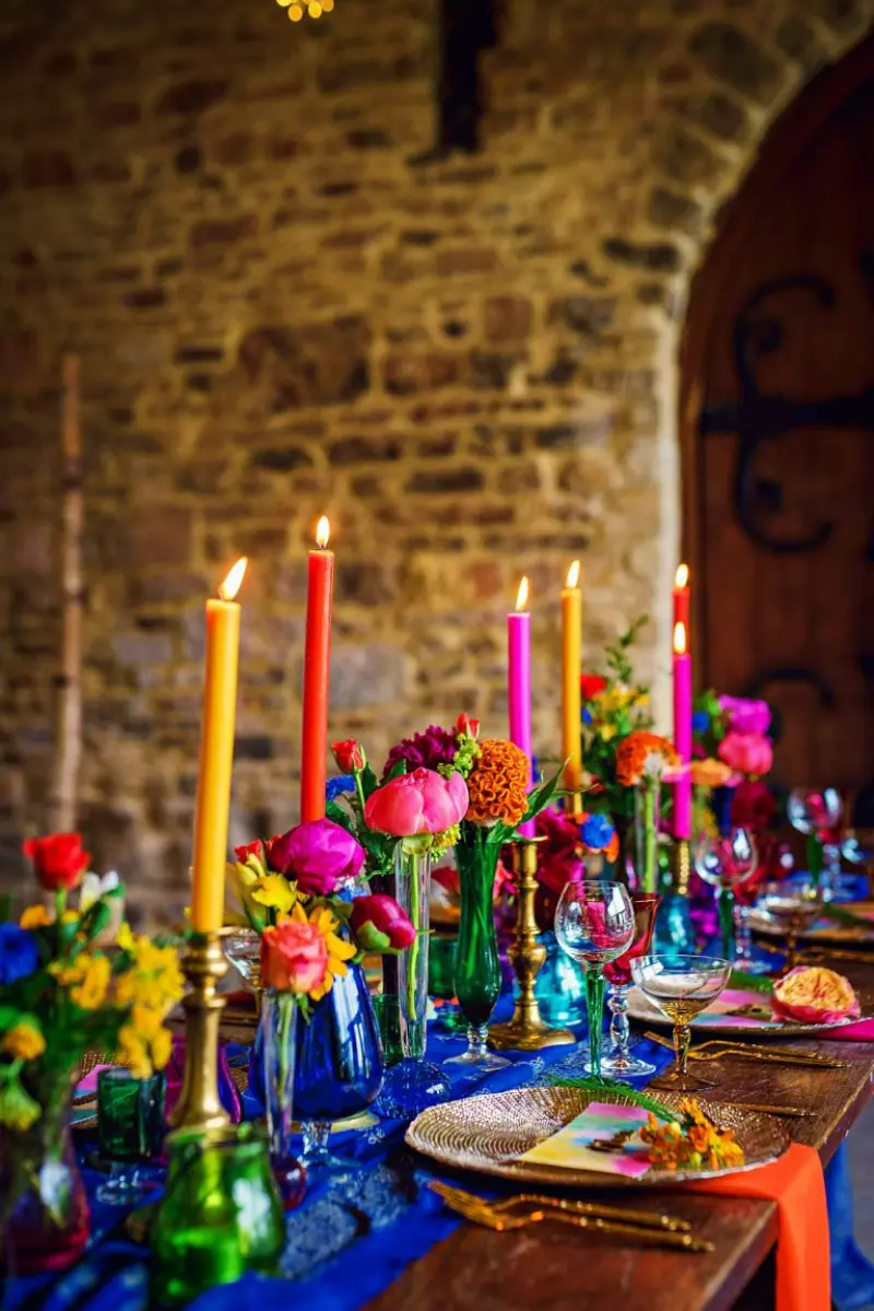 decora&ccedil;&atilde;o colorida para mesa com velas coloridas em casti&ccedil;ais dourados e arranjos de flores tropicais vibrantes.