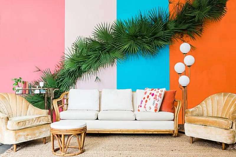 lounge de estilo tropical apresenta uma parede com blocos de cores vibrantes e um sof&aacute; de vime com estofado branco