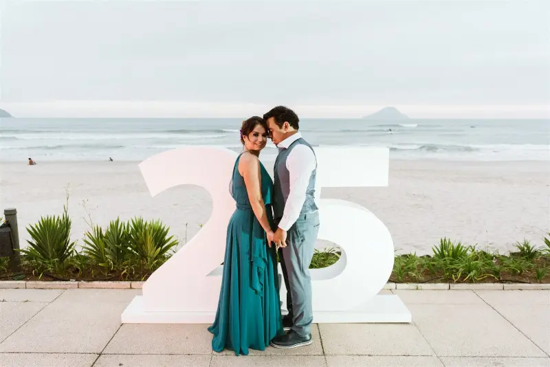 noivos segurando as mãos em frente ao letreiro de 25 anos de casados na praia