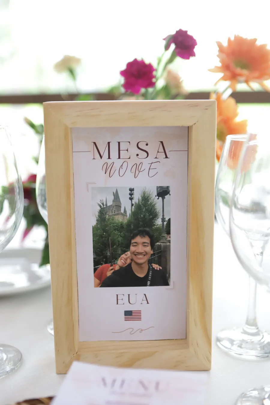 Detalhe da mesa mostrando identificação da mesa com tema de viagem e flores ao fundo.