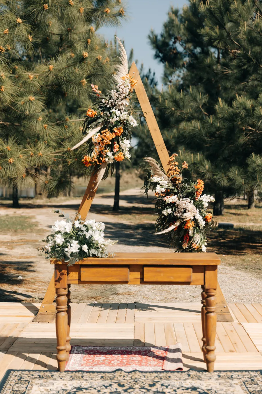 Altar triangular de madeira decorado com flores em tons de terracota, branco e plumas, com uma mesa rústica em frente, para um casamento boho