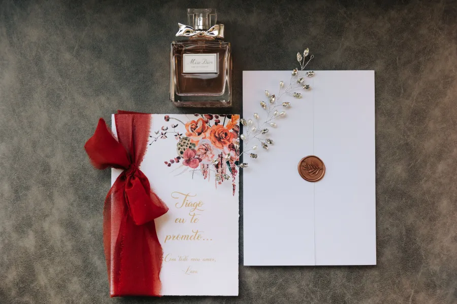Detalhe da papelaria de casamento com convites decorados com flores em tons de terracota e um lacre de cera, ao lado de um perfume e acessório de cabelo