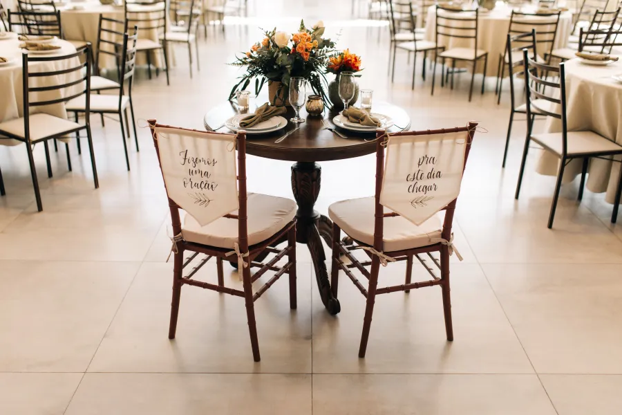 Mesa dos noivos em uma recepção de casamento, decorada com um arranjo floral em tons de terracota e cadeiras com mensagens personalizadas, em um casamento terracota.