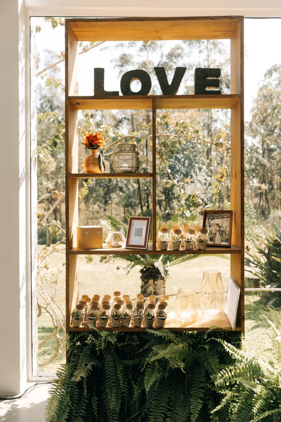 Estante de madeira decorada com o letreiro "LOVE", lembrancinhas de suculentas e fotos, criando um ambiente rústico e charmoso para um casamento boho.