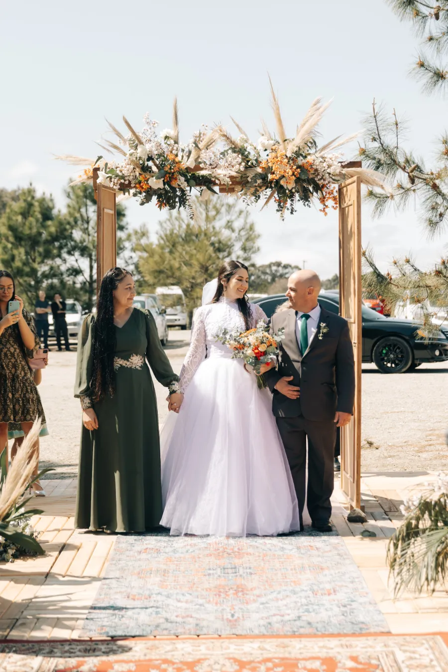 A noiva caminhando pelo corredor com seus pais sob um arco decorado com flores e plumas, em um cenário de casamento boho.