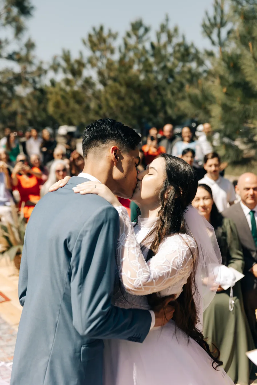 Os noivos se beijando durante a cerimônia ao ar livre, com convidados ao fundo, em um casamento terracota