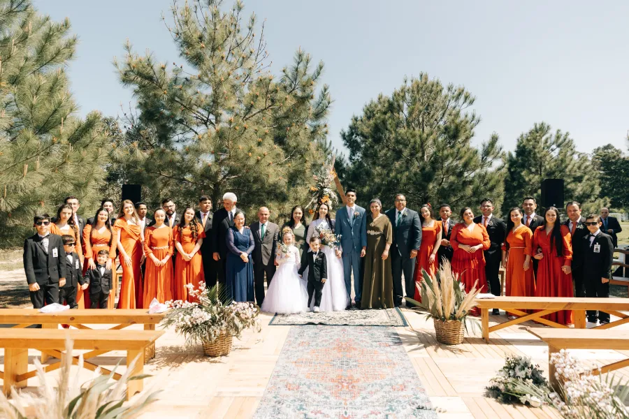 Foto em grupo do cortejo de casamento e convidados em um cenário ao ar livre, com várias madrinhas usando vestidos cor terracota, destacando o tema do casamento terracota