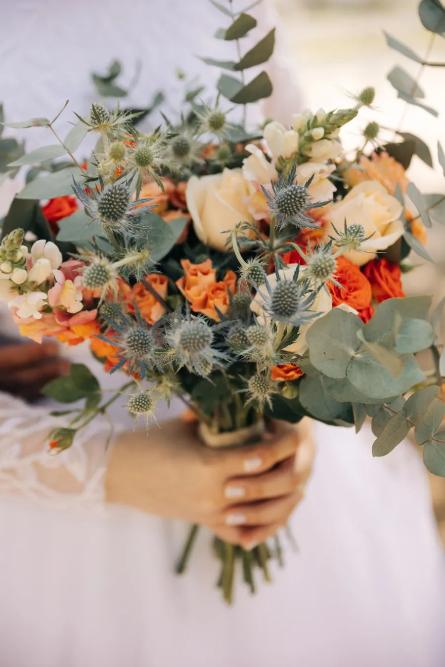 Close-up do buquê da noiva com flores em tons de terracota, branco, folhagens e detalhes azuis, perfeito para um casamento terracota ou boho.