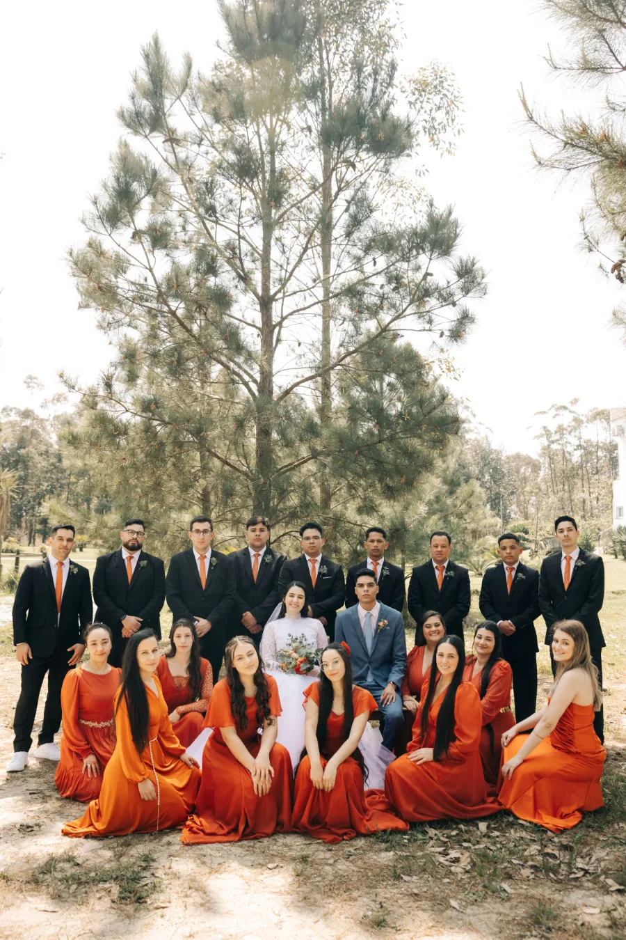 Grupo de padrinhos e madrinhas em um casamento terracota, com os homens usando gravatas laranja e as mulheres vestidos longos cor terracota, posando ao ar livre.