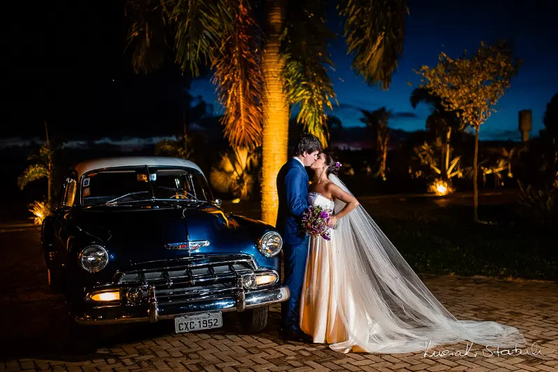 noivos se beijando em frente a carro antigo em casamento à noite