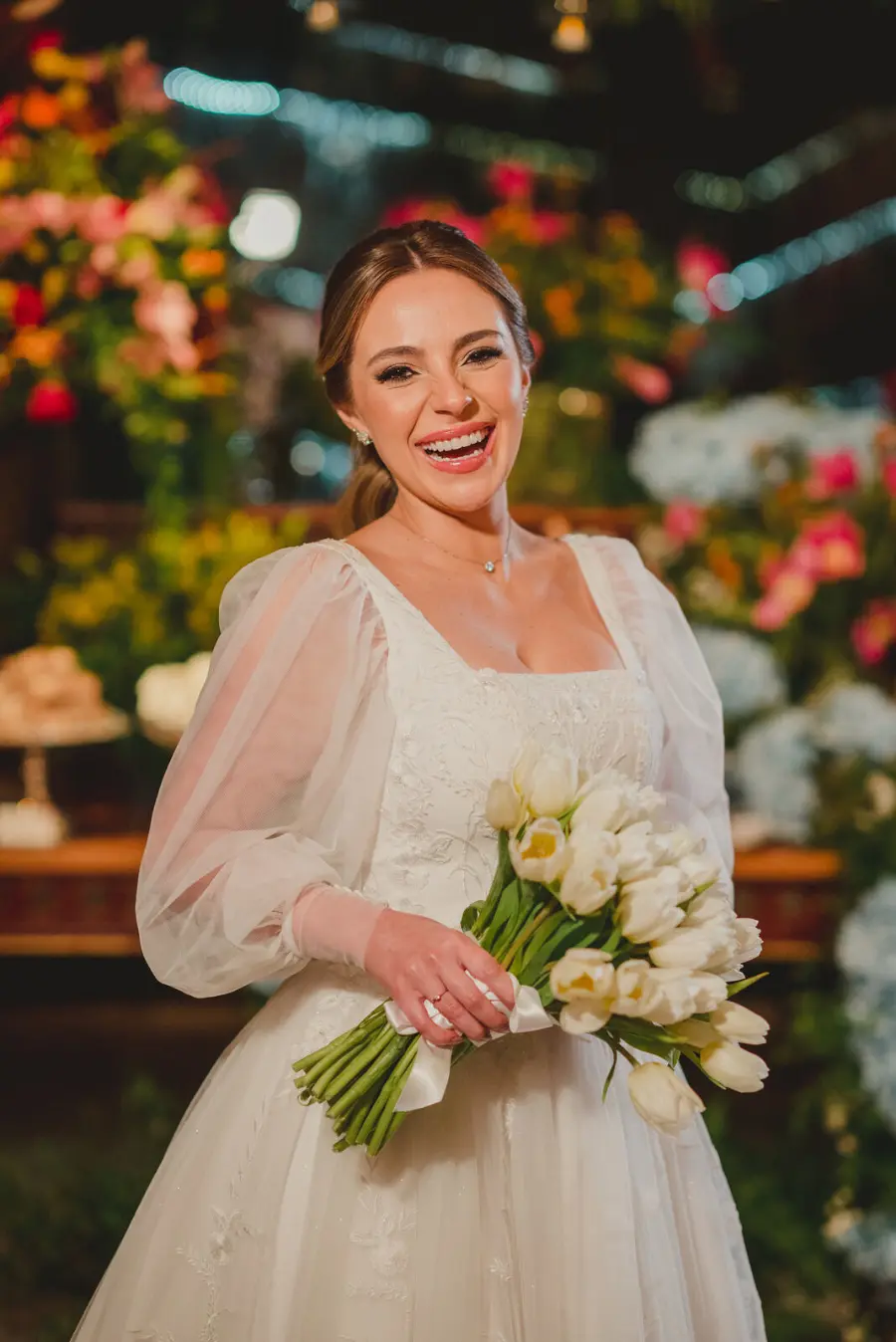 Noiva sorridente com vestido branco de mangas bufantes transparentes, usando colar delicado e segurando buquê de tulipas brancas em ambiente decorado com flores coloridas.