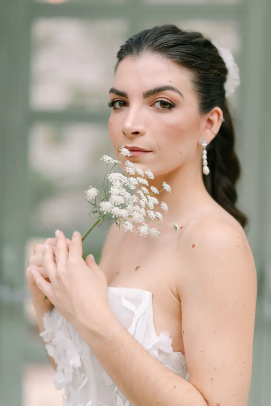 Noiva de perfil com cabelo preso e brincos longos de pérolas, vestindo vestido tomara que caia e segurando flor branca delicada