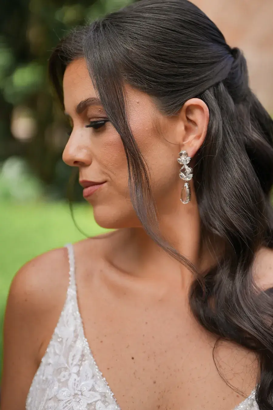 Noiva de perfil com cabelo castanho longo ondulado semi-preso, usando vestido com alças finas e brinco prateado longo com pedras transparentes