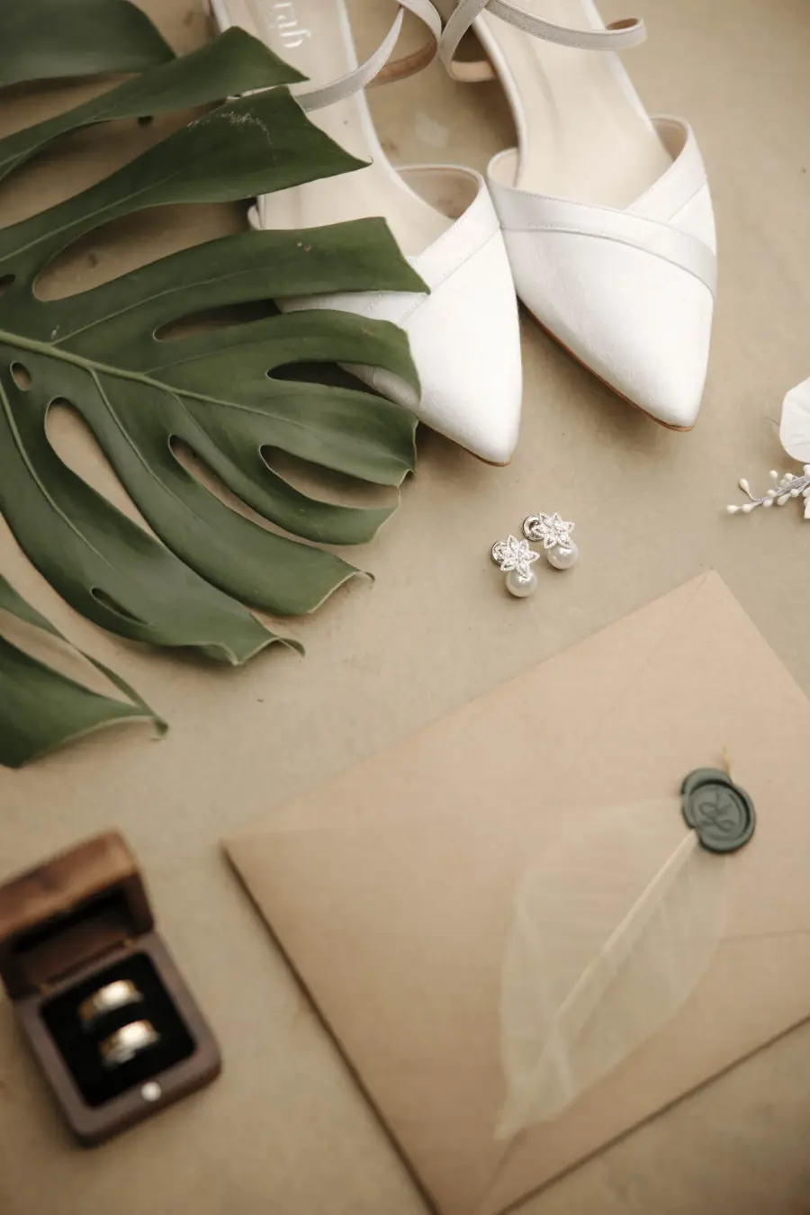 Flat lay de itens de casamento: sapatos brancos de bico fino, folha de costela-de-adão, brincos prateados com pérola, caixa com alianças e envelope bege lacrado com cera e folha esqueleto.