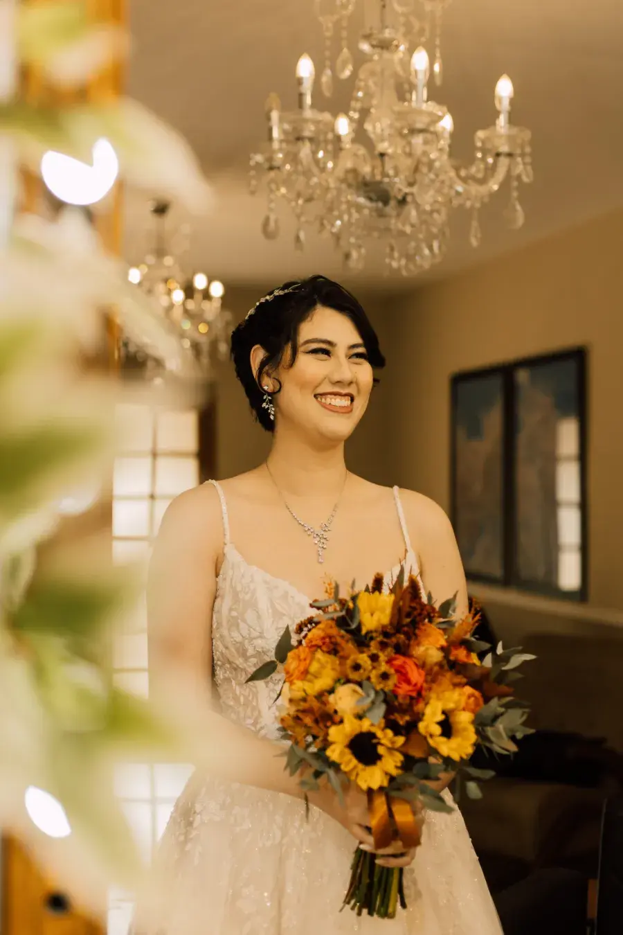 Noiva sorridente com vestido branco de alças finas, usando colar e brincos prateados, segurando buquê de flores amarelas, laranjas e vermelhas em salão iluminado por lustres