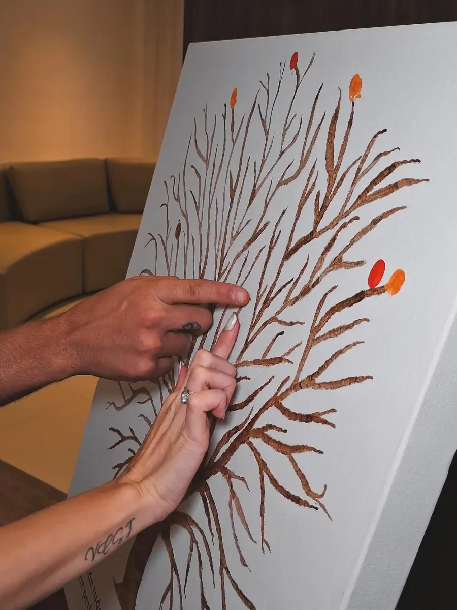 Duas mãos pressionando os dedos pintados em uma tela com galhos, criando folhas coloridas.