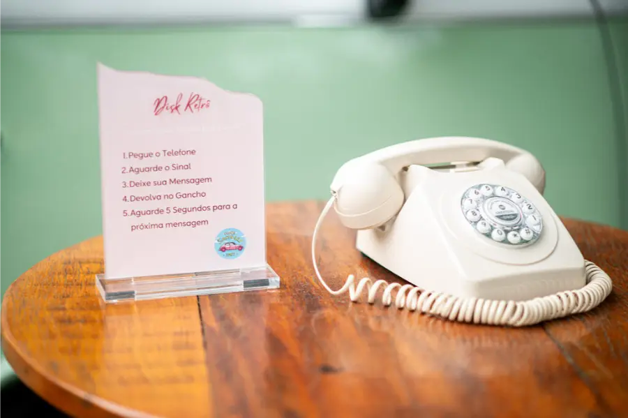Telefone retrô bege em mesa de madeira com plaquinha explicativa ao lado, orientando convidados a deixarem mensagens gravadas durante o casamento
