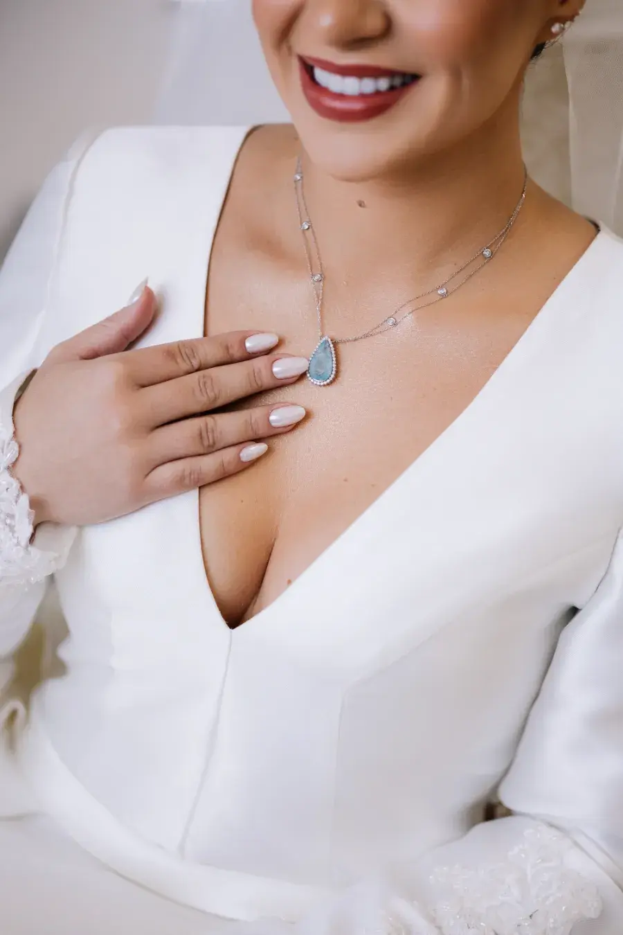 Noiva com vestido branco de decote profundo em V, usando colar prateado com pingente de pedra azul em formato de gota e segurando delicadamente o pingente com a mão.