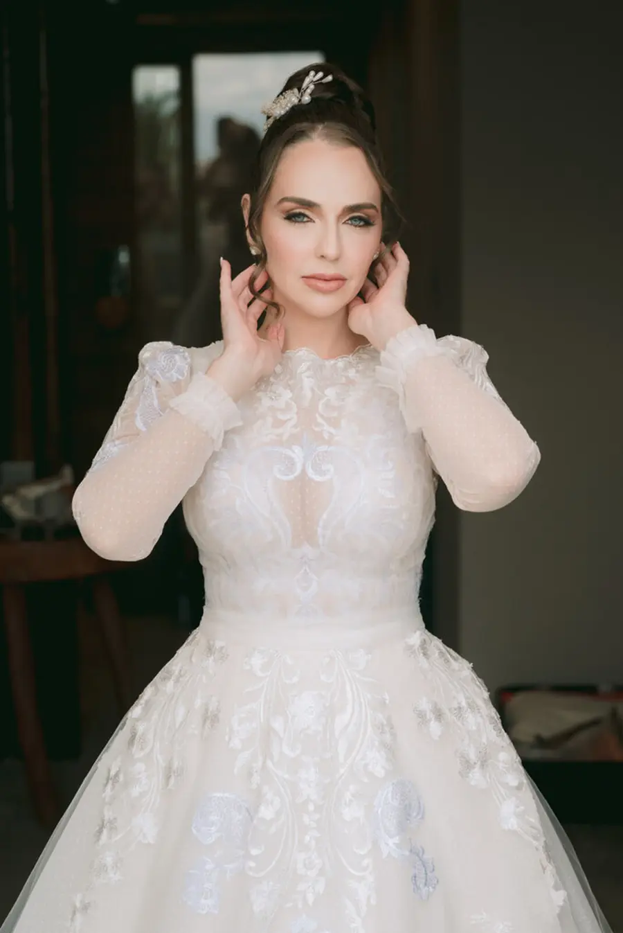 Noiva de frente usando vestido de manga longa com renda e transparência, penteado preso com acessório de cabelo e maquiagem suave