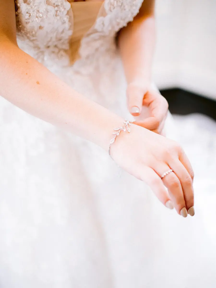 Close das mãos de noiva ajustando pulseira prateada com pingentes delicados, usando vestido branco rendado