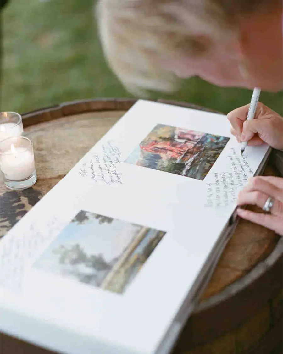 Convidado escrevendo mensagem em um livro de assinaturas com fotos impressas, apoiado em um barril usado como mesa.