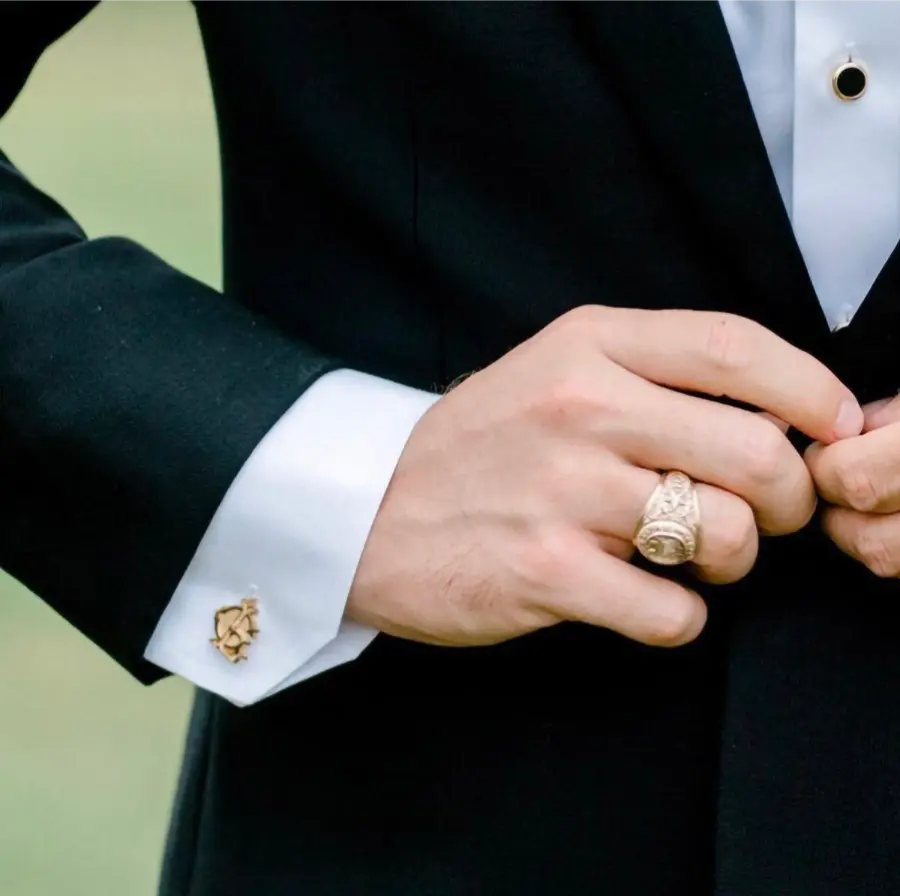 Close do punho de terno preto com abotoadura dourada na camisa branca; mãos ajustando o paletó e anel largo dourado no dedo