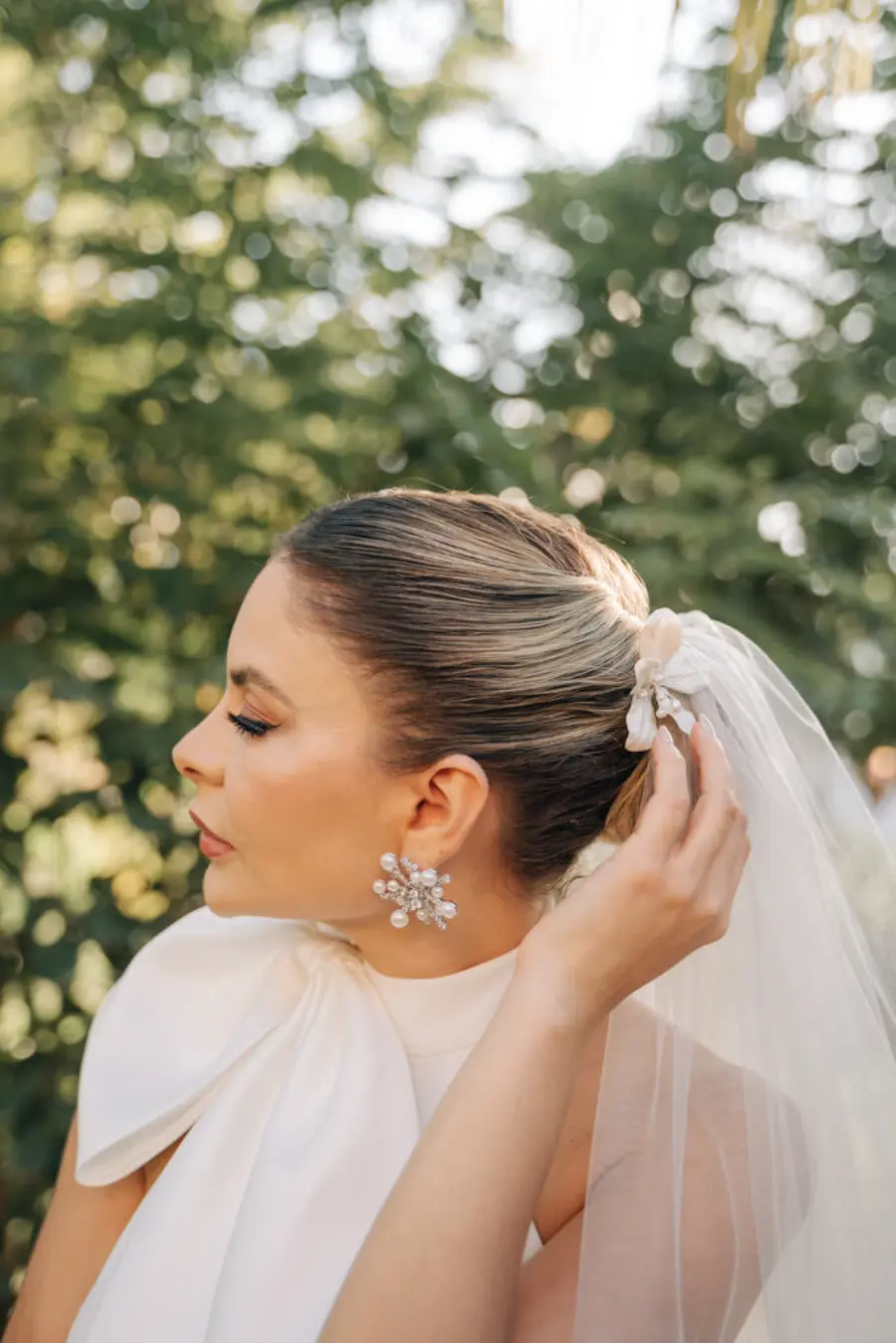 Noiva de perfil com cabelo preso em coque alto, usando véu preso com laço de tecido e brinco prateado em formato de flor com pérolas
