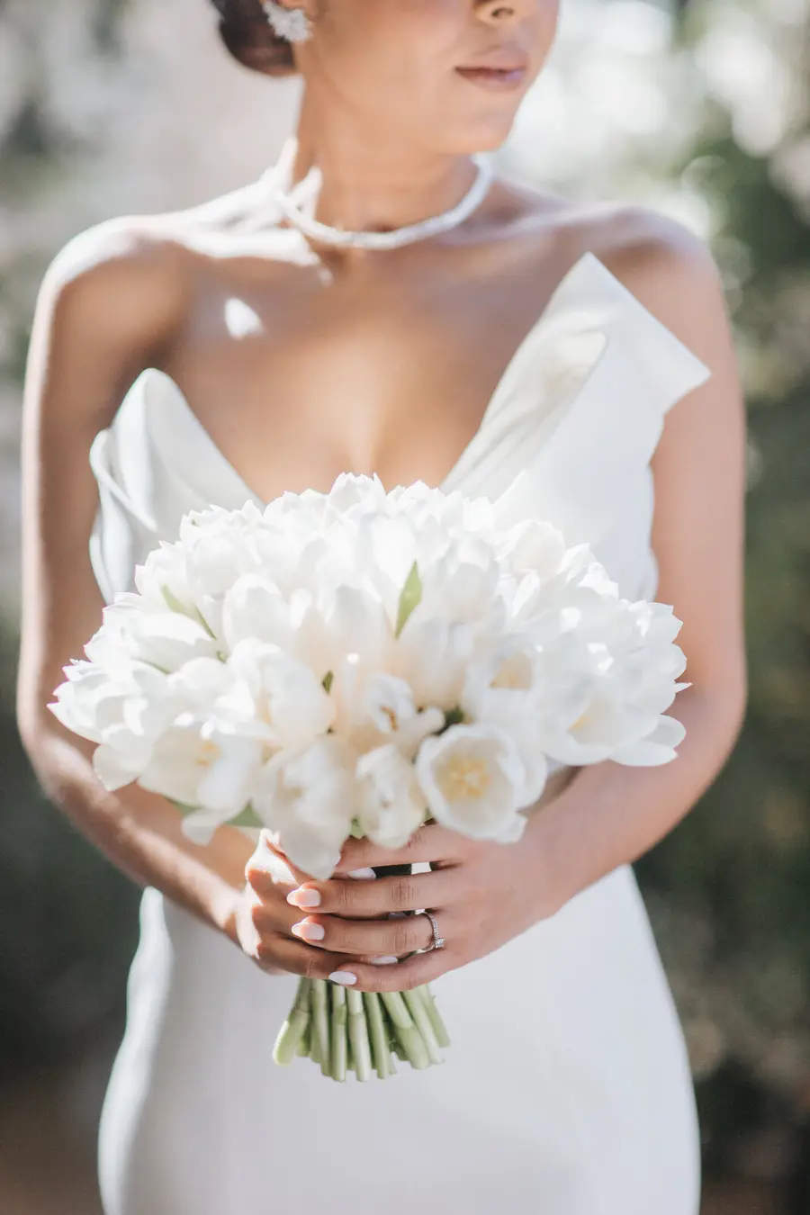 Noiva segurando buquê de tulipas brancas, usando vestido branco com decote profundo estruturado e colar de pérolas