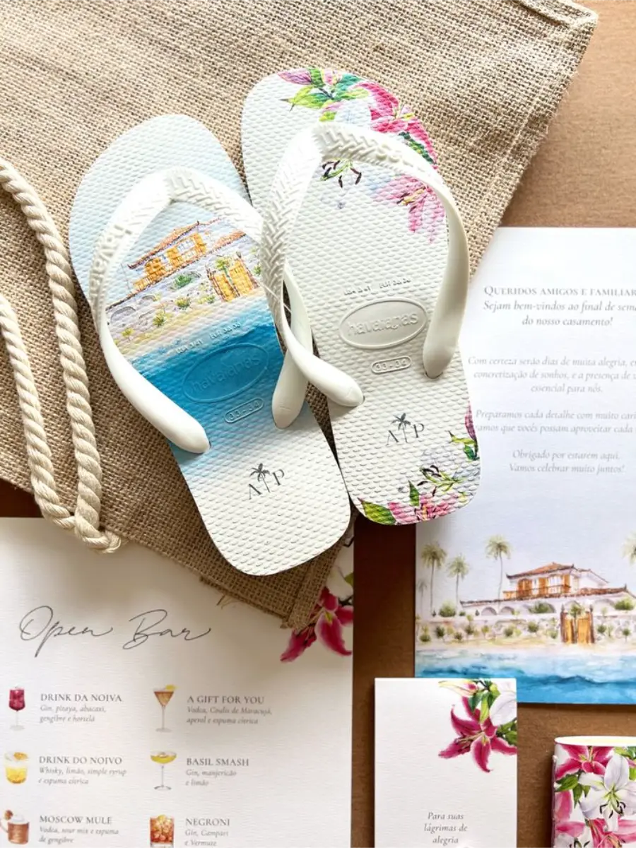 Chinelos personalizados brancos com estampa floral e paisagem litorânea, acompanhando identidade visual tropical do casamento.