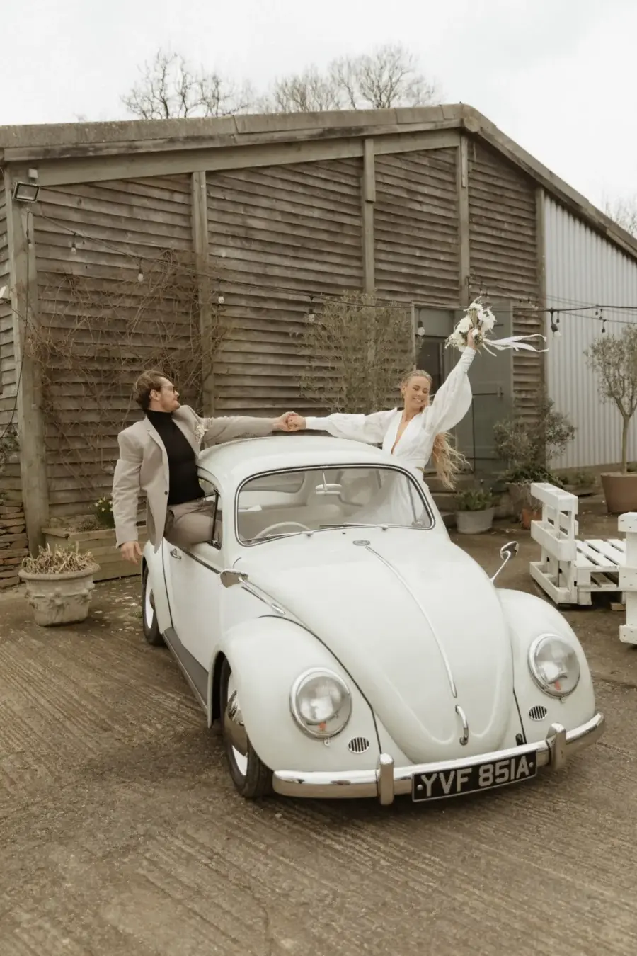 Noivos celebrando sobre Fusca branco antigo estacionado em frente a galpão rústico.