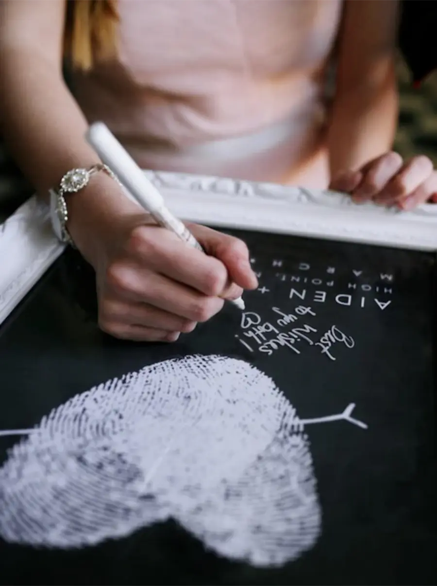 Convidada escrevendo mensagem em quadro de assinatura com impressão digital em formato de coração para casamento.