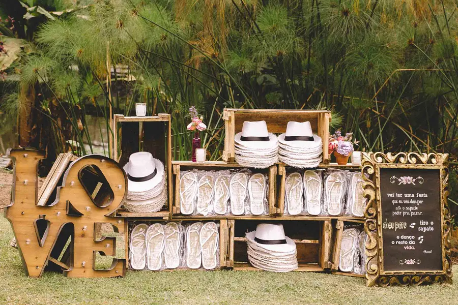 Expositor rústico com chinelos personalizados brancos e chapéus em casamento ao ar livre.