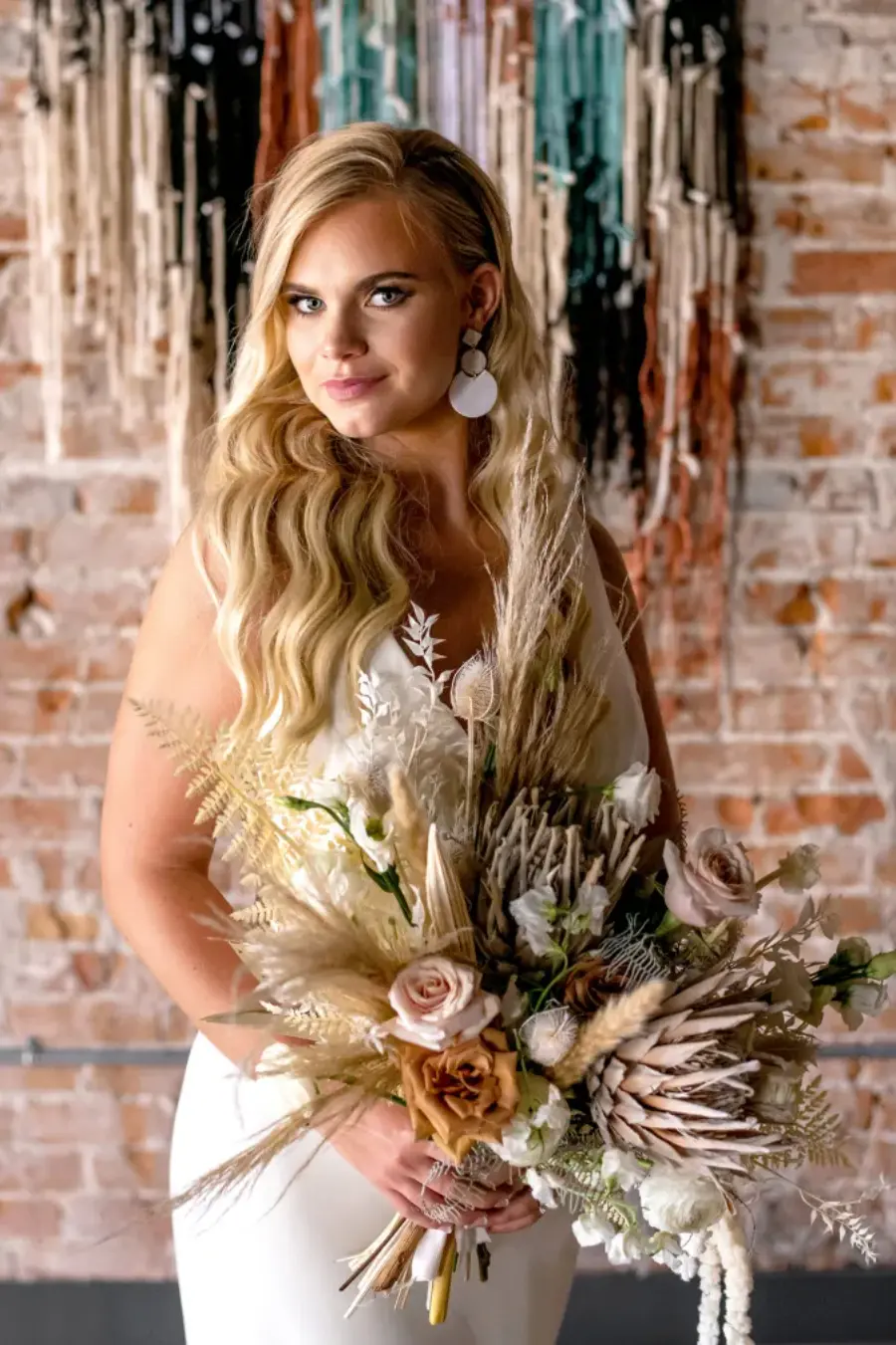 Noiva loira com cabelo ondulado solto, usando vestido branco sem mangas, brincos brancos redondos e segurando buquê com flores secas e rosas em tons terrosos