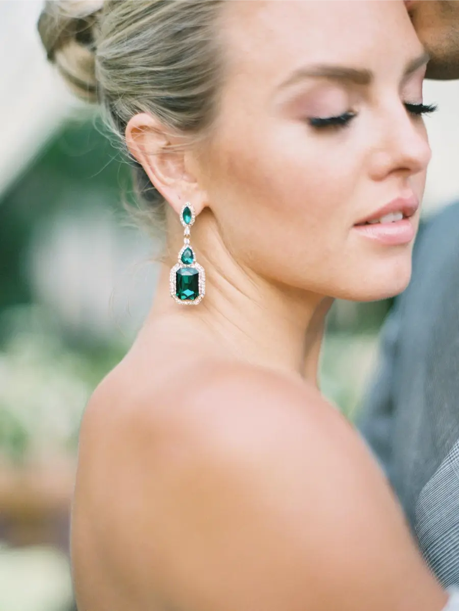 Noiva de cabelo preso, usando brinco longo com pedras verdes e detalhes em diamantes, em momento íntimo com parceiro.