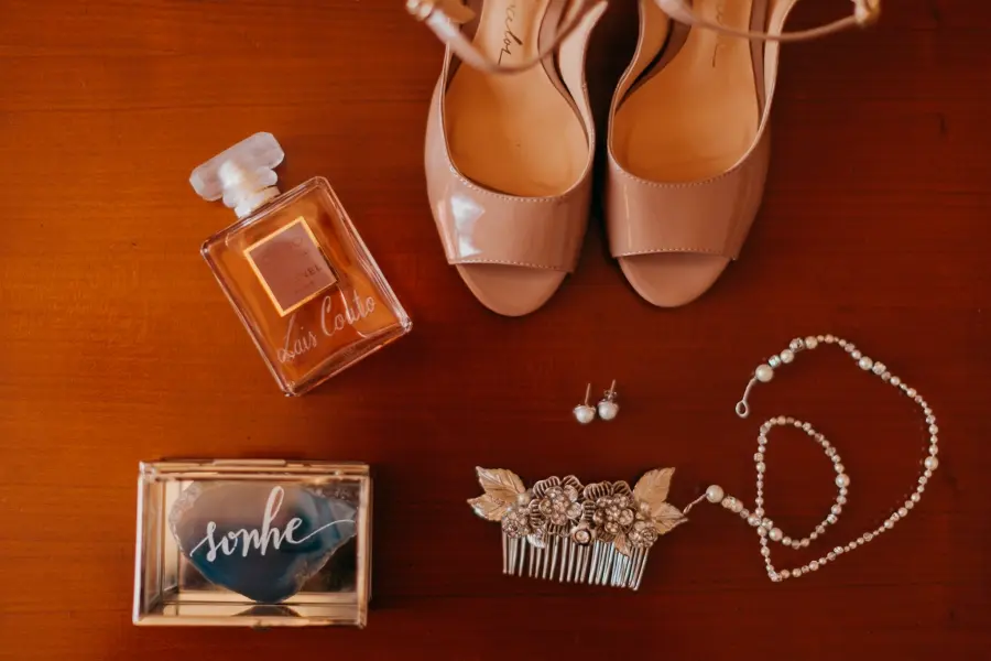 Conjunto de acessórios de noiva sobre superfície de madeira, incluindo sapatos nude de salto, perfume, caixa com a palavra 'sonhe', par de brincos de pérola, presilha floral prateada e colar de pérolas.